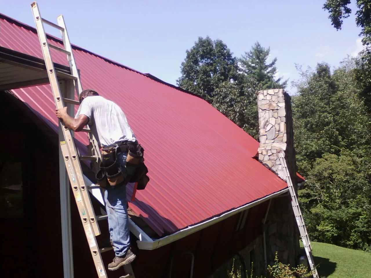 Roofer on a ladder beside a red metal roof and stone chimney — Soffit & Fascia Repair by Roofing Brothers in Chambersburg